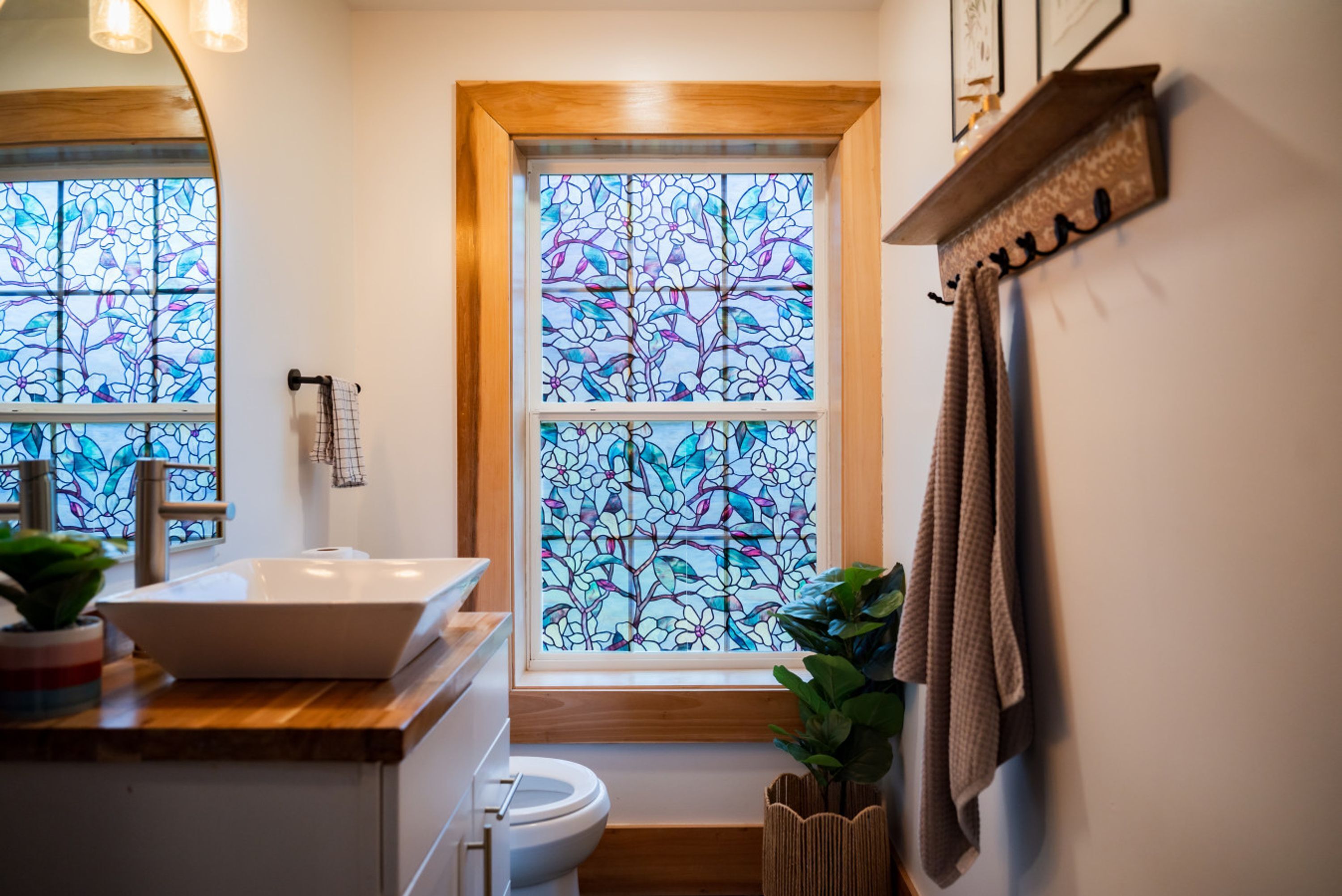 A bathroom with a wooden vanity, a white sink, a glass-encased window with a floral pattern, and a towel rack.