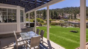 A covered patio with a glass table and wicker chairs, overlooking a well-maintained lawn and a lake with houses in the background. - Lakefront Big Bear in Big Bear Lake (Big Bear Lake)