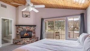 A bedroom with a stone fireplace, a large bed, a ceiling fan, and a balcony with a view of trees. - Lakefront Big Bear in Big Bear Lake (Big Bear Lake)