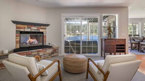 A living room with a stone fireplace, white leather chairs, a woven ottoman, and a view through large glass doors to a deck. - Lakefront Big Bear in Big Bear Lake (Big Bear Lake)