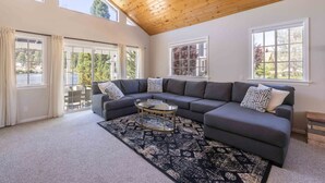 A spacious living room with a large sectional sofa, a glass coffee table, and a patterned rug. There are large windows providing natural light and views of the outdoors. - Lakefront Big Bear in Big Bear Lake (Big Bear Lake)