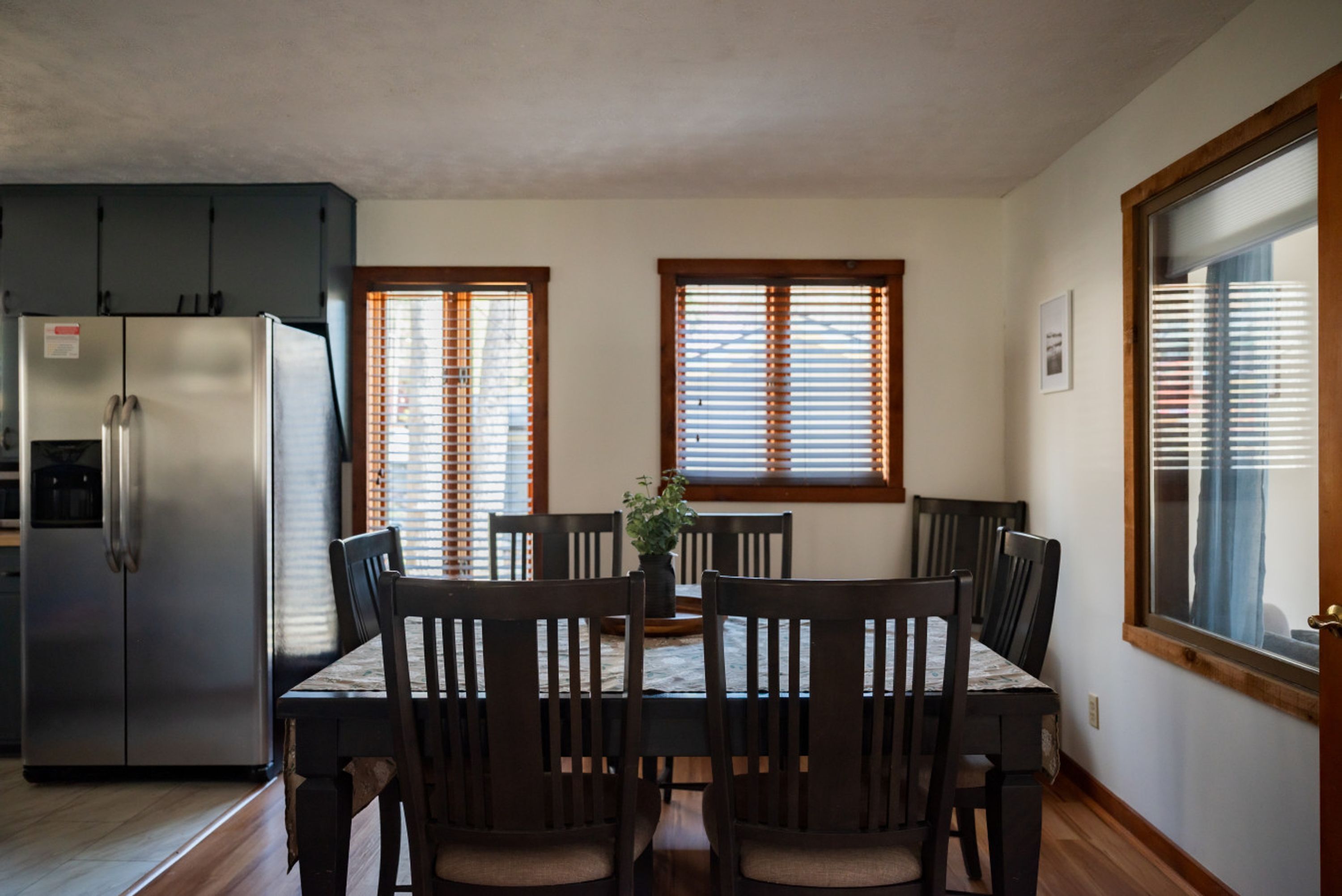 A kitchen with a refrigerator, dining table, and chairs. There are windows with blinds and a view of the outside.
