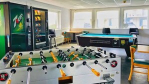 Une table de baby-foot, une table de billard et un distributeur de boissons 7UP dans une pièce dotée de grandes fenêtres.