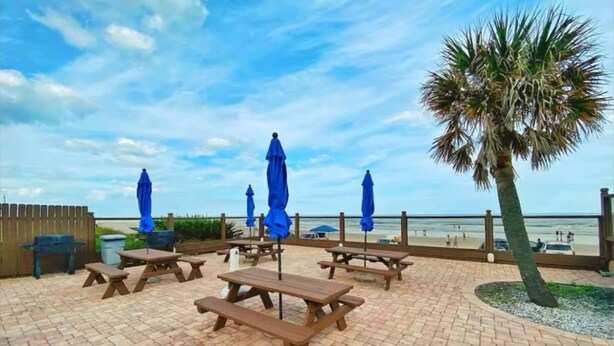 Une terrasse extérieure avec des tables de pique-nique et des parasols bleus, donnant sur une plage.
