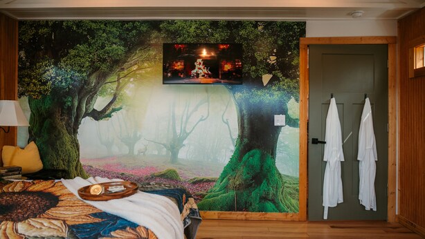 Una habitación con un gran mural de un bosque, una cama con colcha estampada de flores, un televisor montado en la pared y una puerta de madera con toallas colgadas.