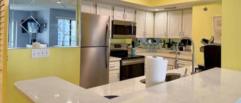 A modern kitchen with a stainless steel refrigerator, microwave, and oven. There is a marble countertop and a sink with a faucet. The kitchen has white cabinets and a yellow accent wall.