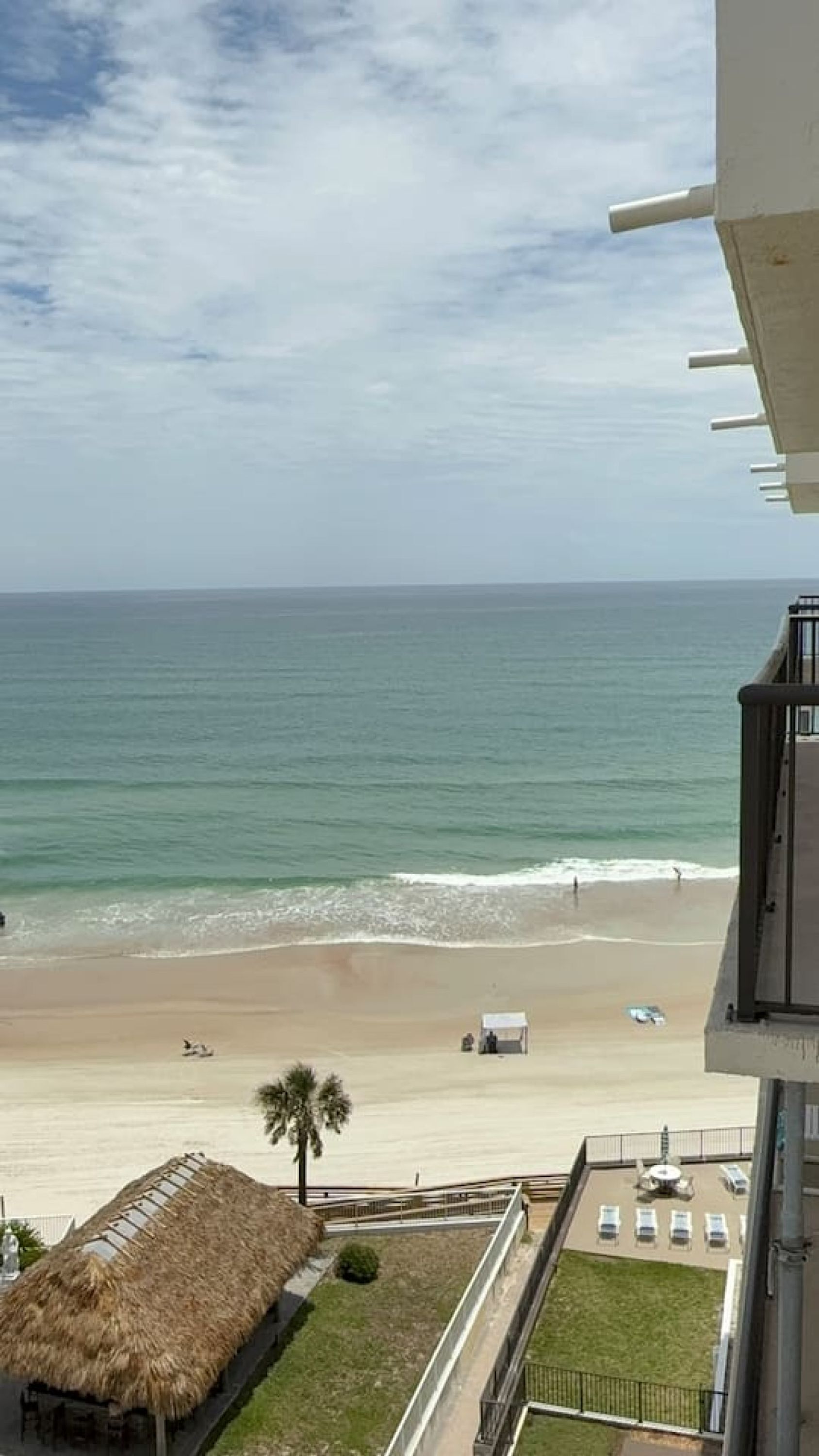 A beach view from a balcony with a thatched-roof structure, a grassy area, and a parking lot.