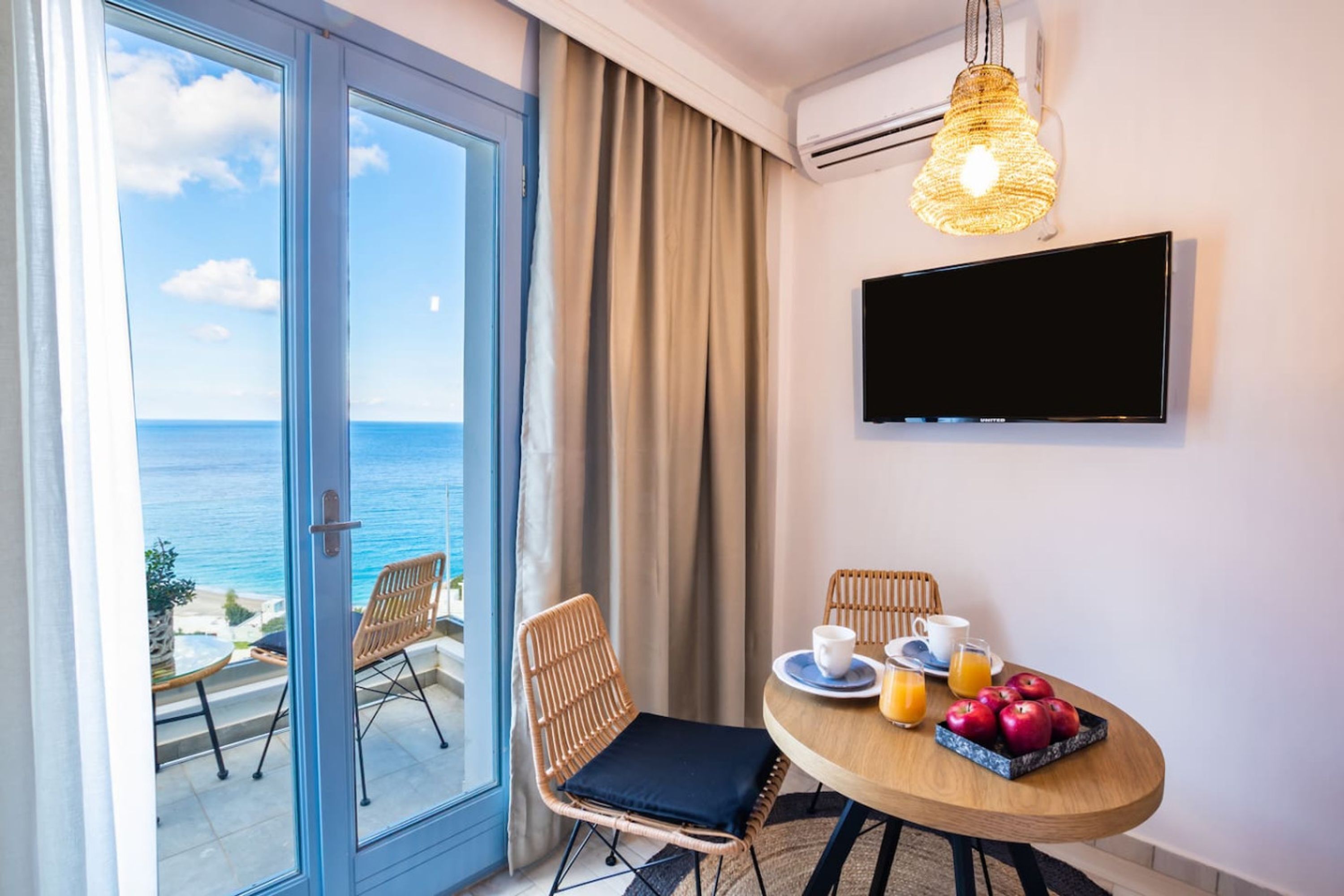 A room with a balcony, a dining table with fruits and beverages, and a TV mounted on the wall.