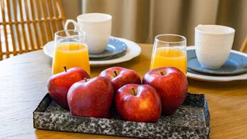 Ein Frühstücksstillleben mit Äpfeln und Orangensaft auf einem Tablett, zwei weiße Tassen und ein Korbstuhl im Hintergrund.