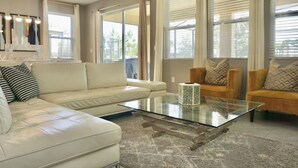 A modern living room with a glass coffee table, beige sofas, and large windows with curtains.