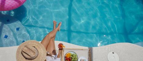 Une vue sur la piscine avec une personne se relaxant, un flotteur rose et un plateau avec de la nourriture et des boissons.