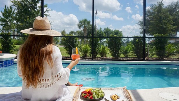 A person relaxing by a pool with a tray of fruits and a drink, overlooking a clear blue pool and greenery.