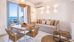 A modern living room with a wooden dining table, wicker chairs, a beige sofa with patterned cushions, and a view of the sea through a large window. - Yploui Suites 3 (Karpathos)