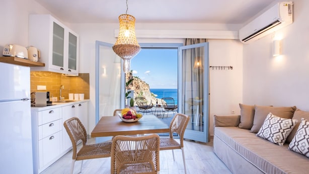 A modern kitchen with white cabinets, a dining table with wicker chairs, and a view of the sea through an open door. - Yploui Suites 3 (Karpathos)