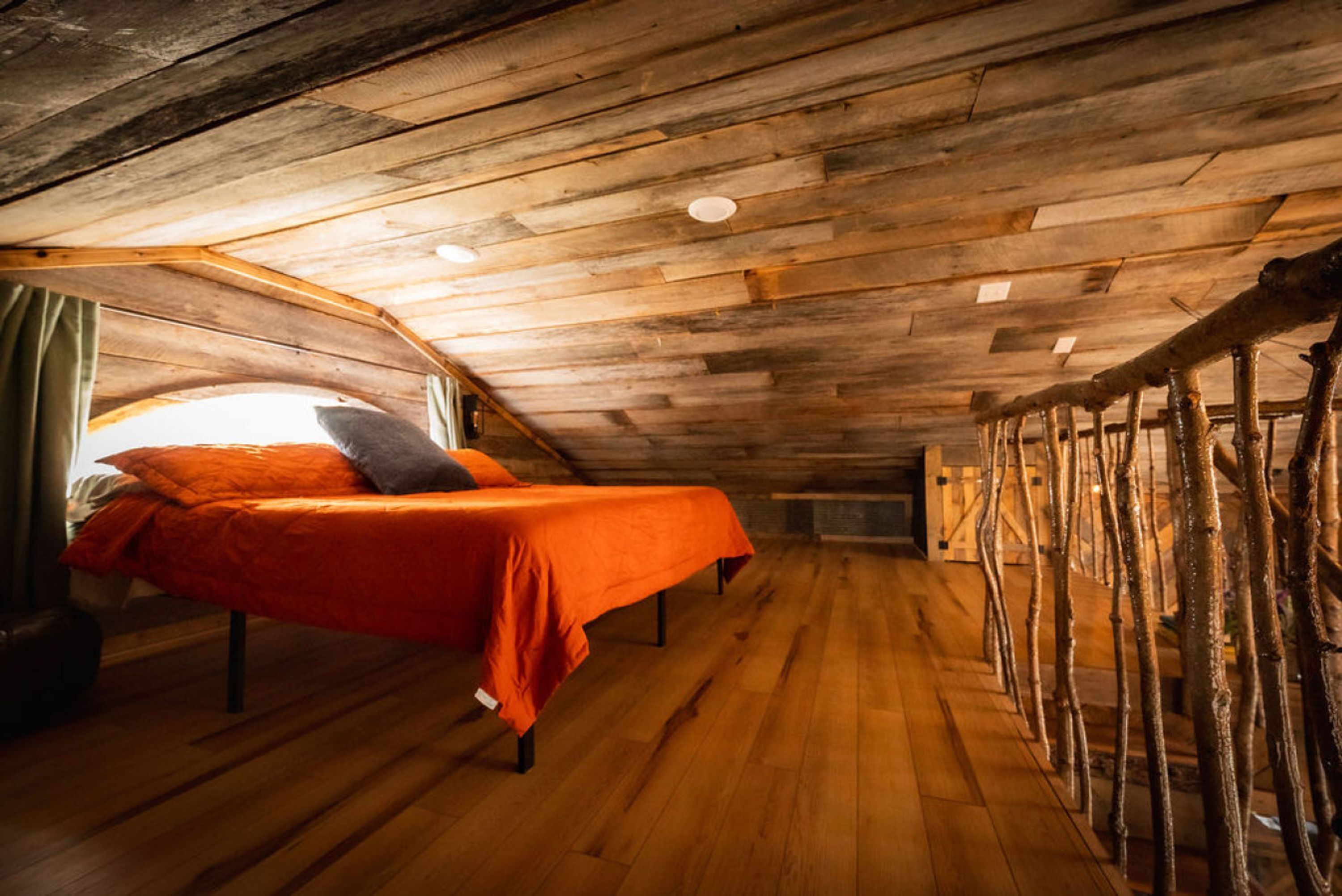 Dormitorio tipo loft con techo inclinado, vigas de madera y cama con colcha naranja.