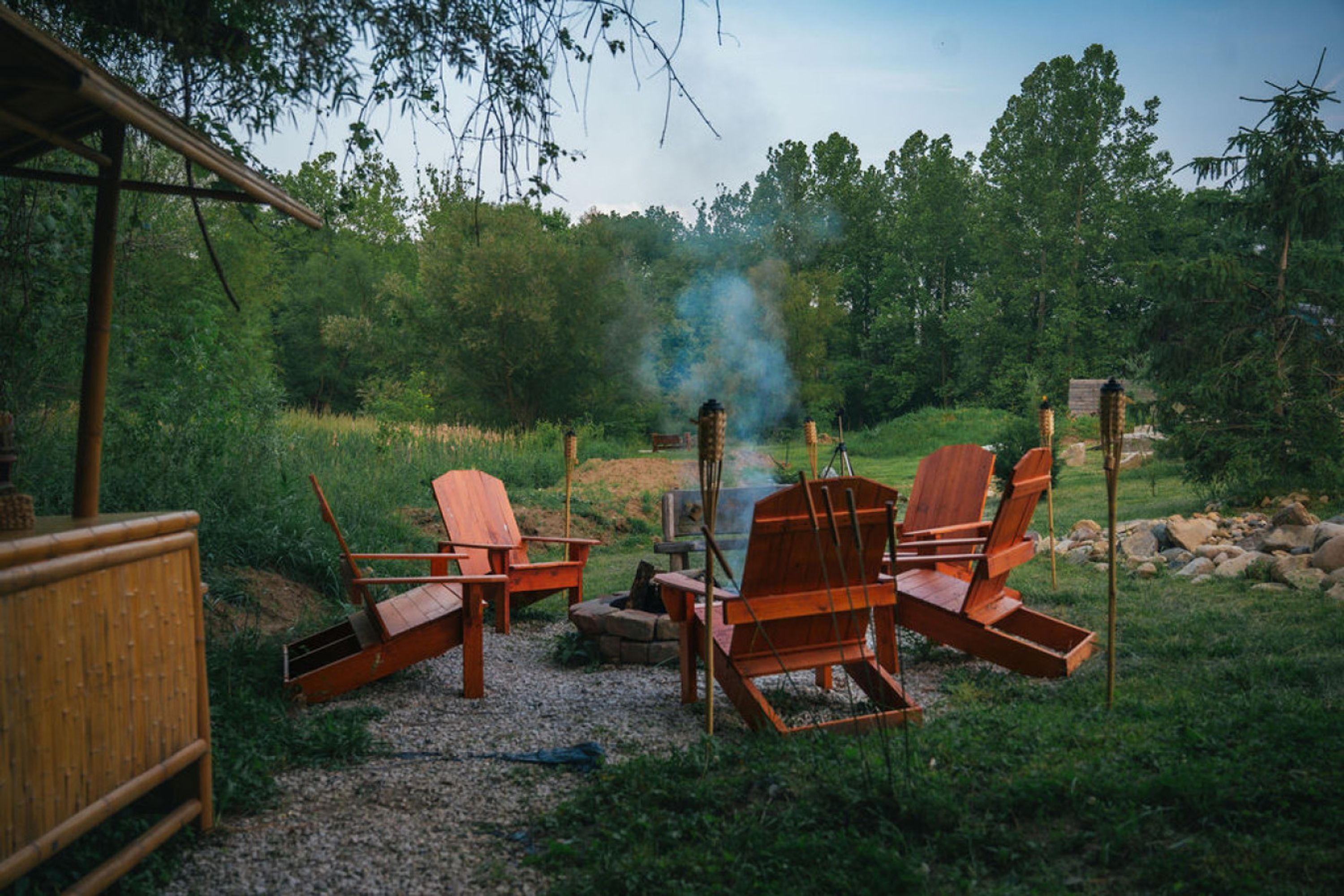 A rustic outdoor seating area with wooden chairs and a fire pit surrounded by greenery.