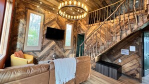 Interior acogedor de una cabaña con suelo de madera en espiga, un sofá de cuero marrón, un televisor de pantalla plana montado, una escalera de madera y una lámpara circular.