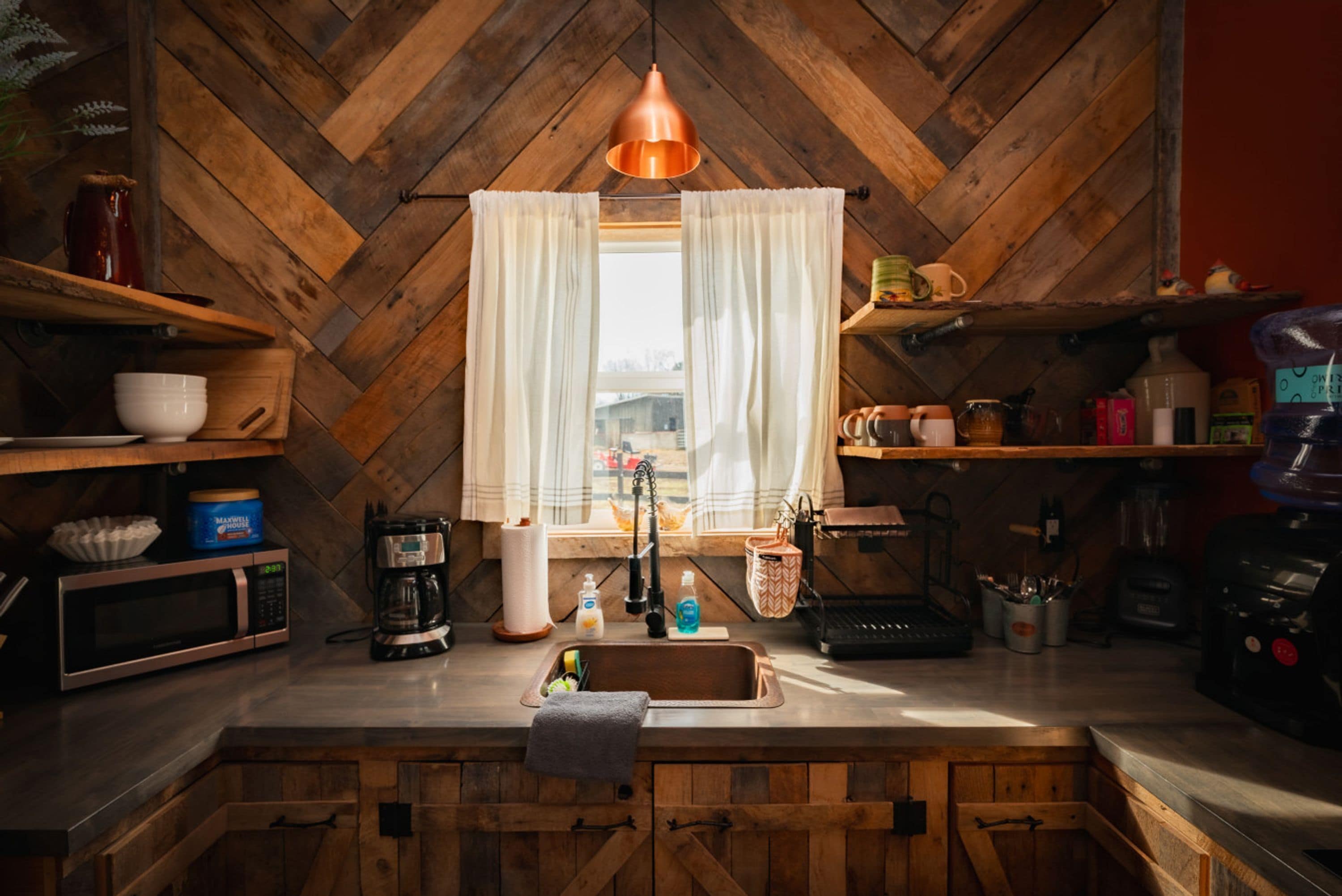 Een keuken met houten visgraatvloer, een raam met witte gordijnen, een koperen hanglamp, en diverse keukenapparaten en keukengerei.