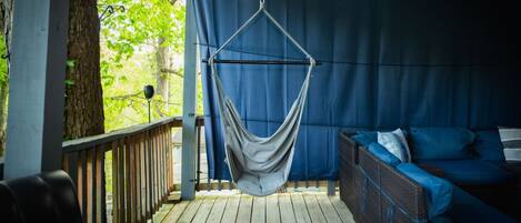 A hammock suspended from a ceiling in a covered outdoor area with a wooden deck, a blue sofa, and a view of trees.