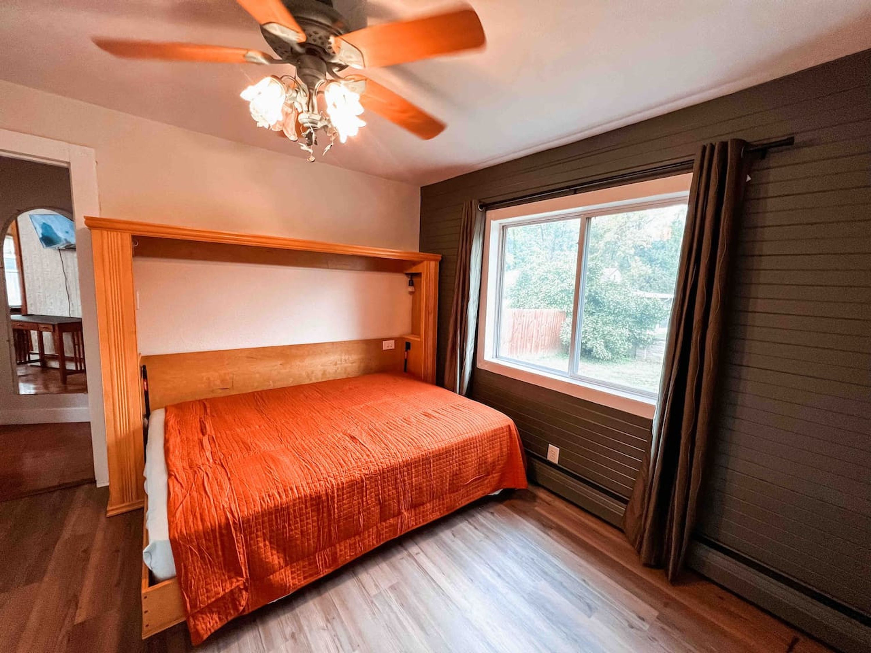 A bedroom with a wooden headboard, a single bed with an orange bedspread, a ceiling fan, and a window with curtains.