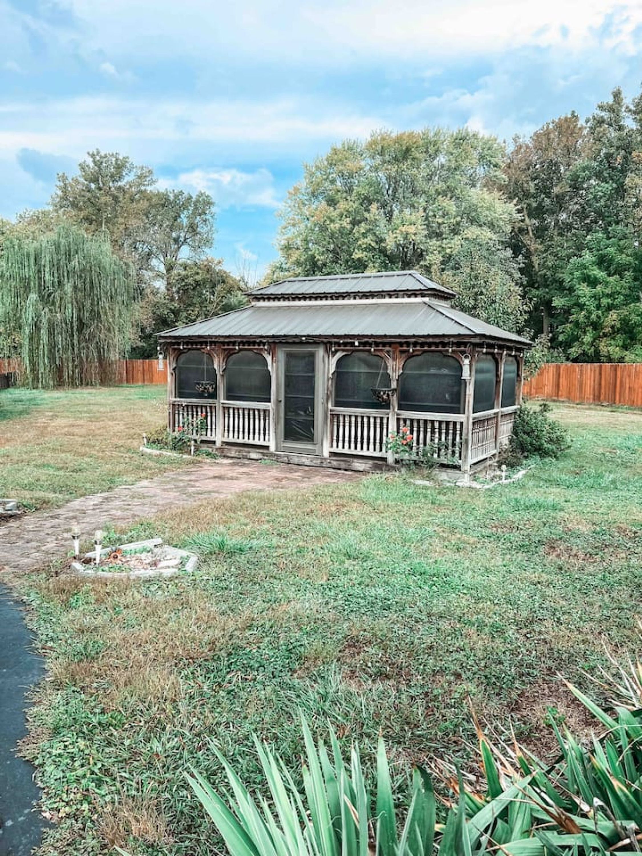 A small wooden gazebo with a porch, surrounded by a grassy yard and trees.