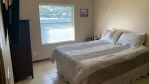 Ein Schlafzimmer mit Bett, Nachttisch, Fenster mit Jalousien, Deckenventilator und einem an der Wand hängenden Bild.