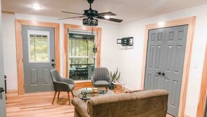 A living room with a sofa, two armchairs, a coffee table, and a ceiling fan. There are two doors and a window with a view of the outdoors. - Two Lakefront Cabins Next Door (Martinsville)