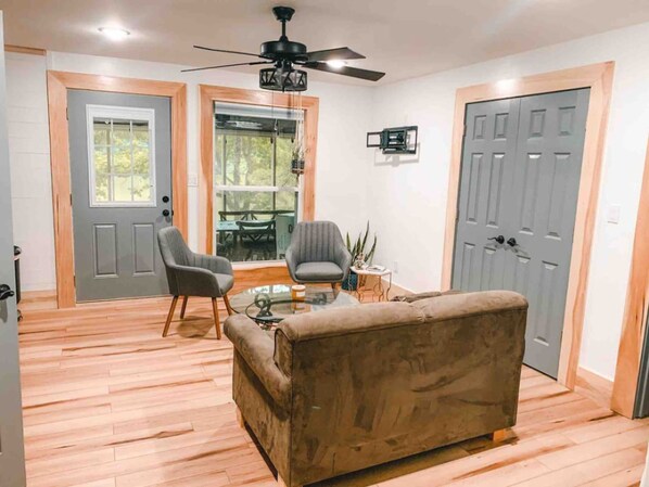 Un salón con sofá, dos sillones, una mesa de centro y un ventilador de techo. Hay dos puertas y una ventana con vistas al exterior.