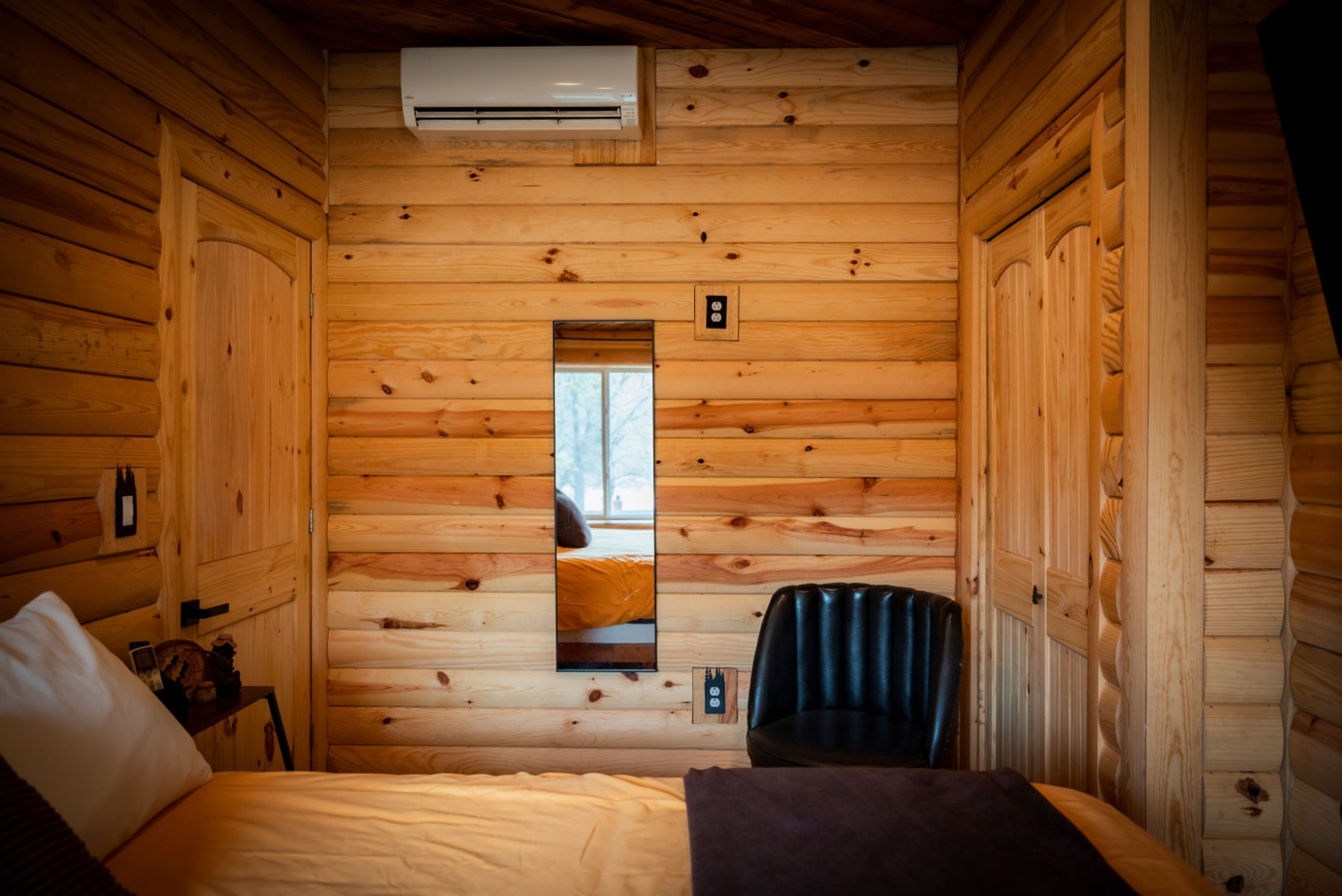 A wooden cabin room with a bed, a chair, an air conditioning unit, and a mirror on the wall.