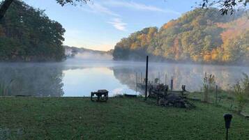 A serene lakeside view with a wooden deck, a rustic fire pit, and a bench.