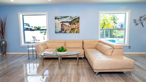 A modern living room with a beige sectional sofa, a glass coffee table with a plant, and large windows offering a view of the outdoors. - Fun Pool Relax16miles Bushgarden (Tampa)