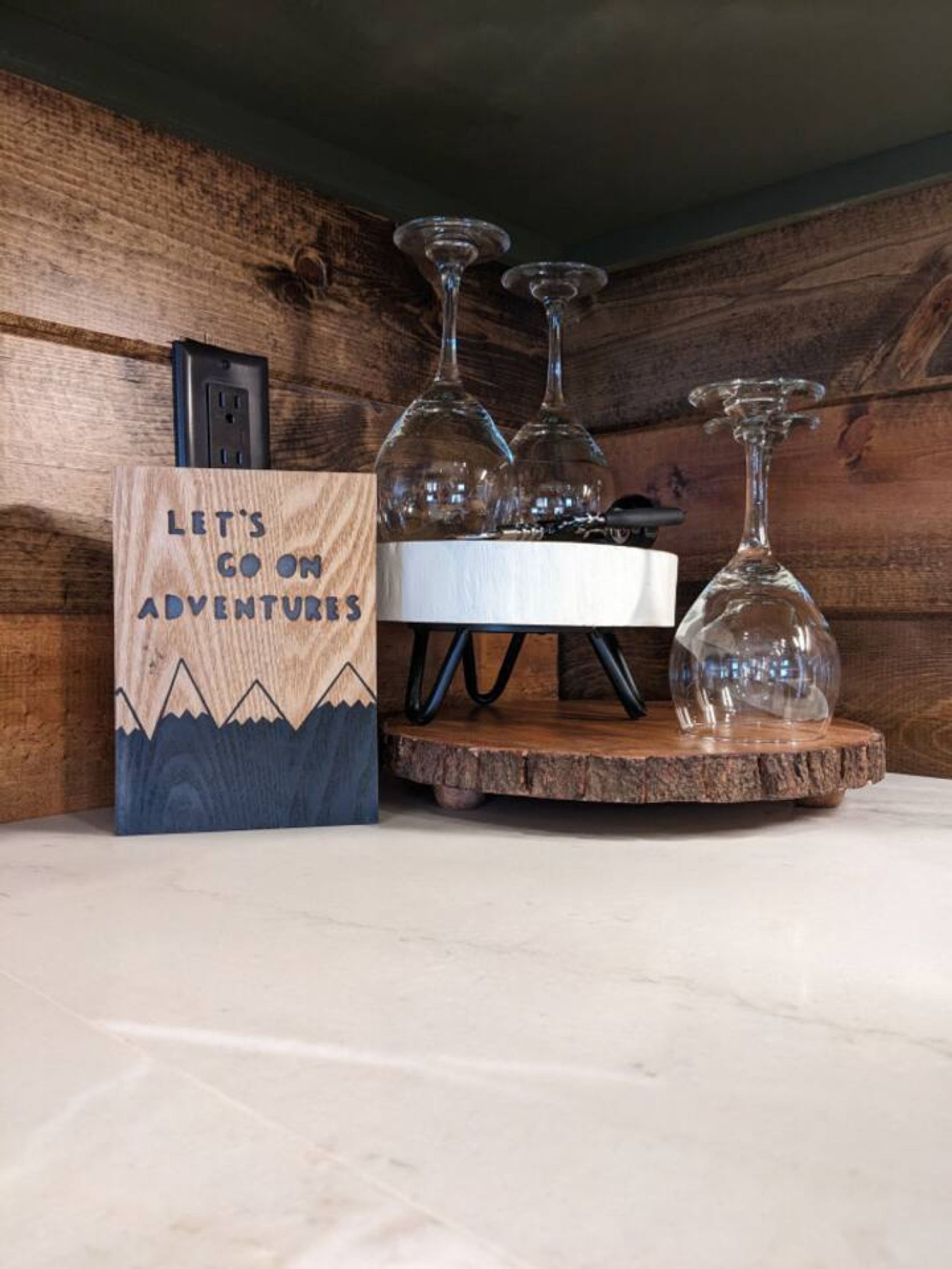 Un letrero de madera con un diseño de montaña y el texto "LET'S GO ON ADVENTURES", junto con tres copas de vino sobre un posavasos de madera.