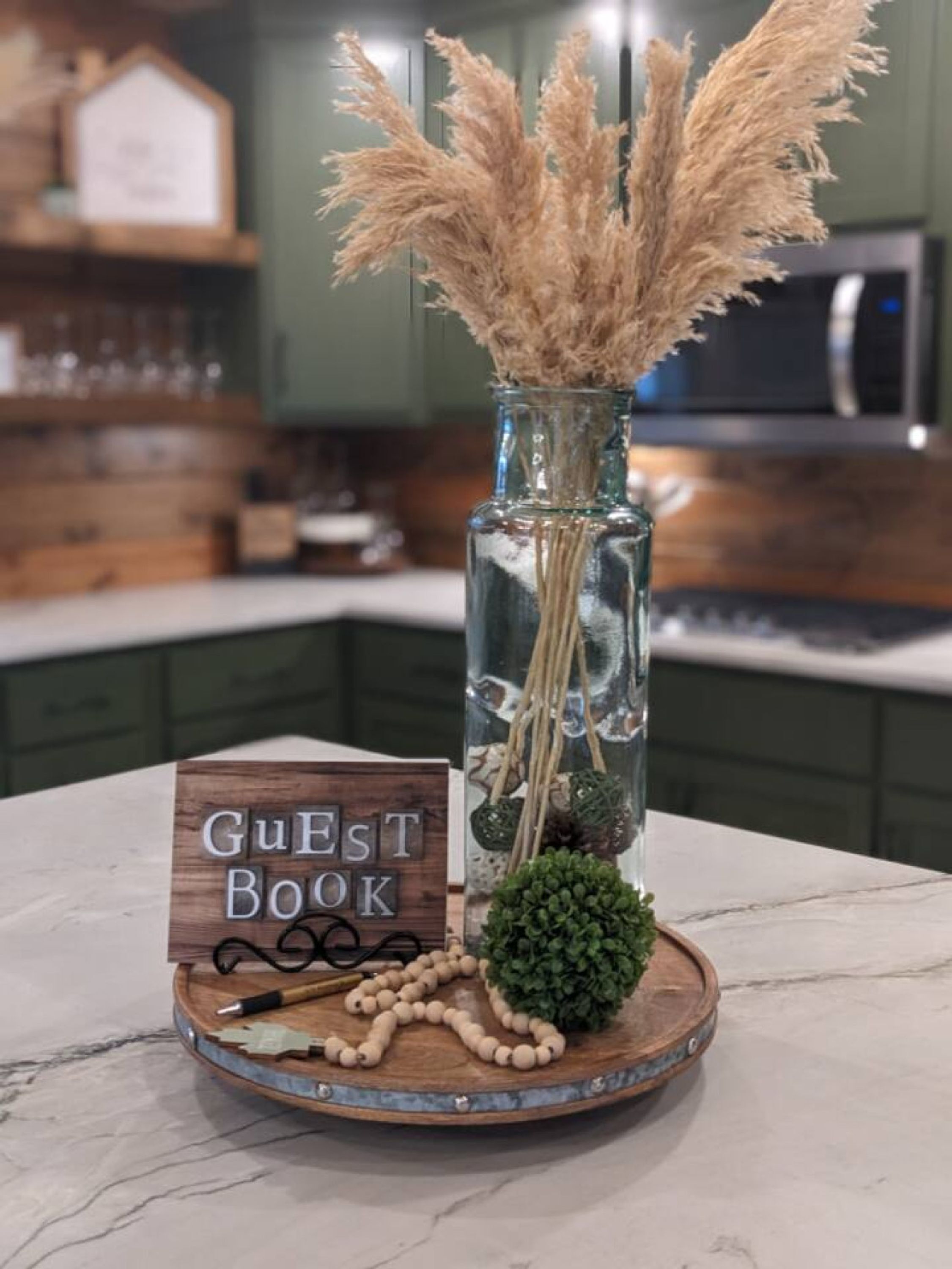Un jarrón de vidrio con plumas de pampa y una pequeña planta verde sobre un posavasos de madera, una señal de madera que dice “Libro de visitas”, y una guirnalda de cuentas blancas sobre el mismo posavasos.