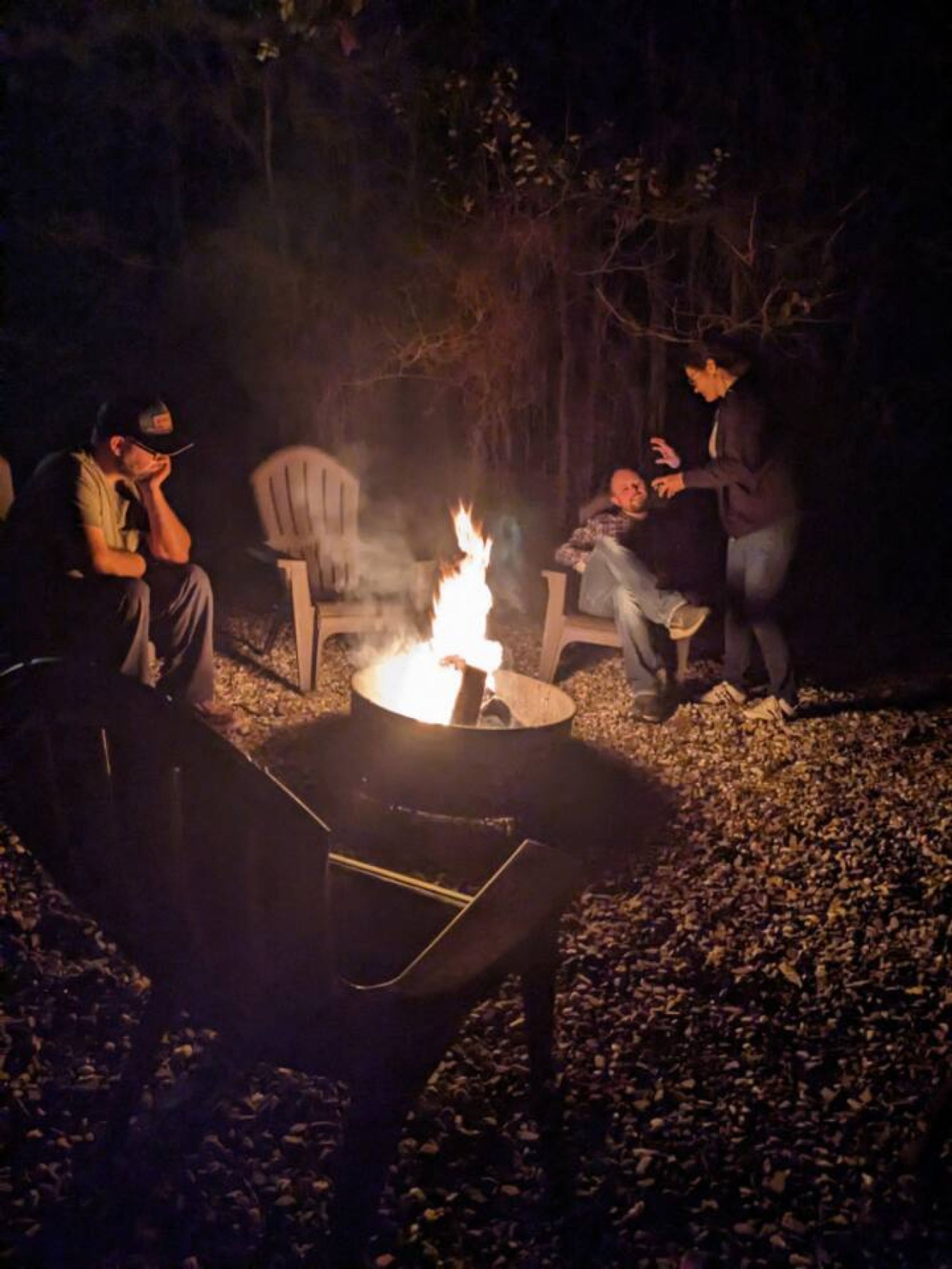 Un grupo de personas alrededor de una fogata en un entorno al aire libre, con sillas y un brasero.