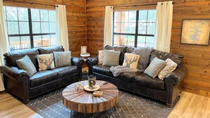 A cozy log cabin interior with two leather sofas, a round coffee table, and large windows with curtains. - Lofted Pines Hot Tub Arcade Air Hockey (Broken Bow)