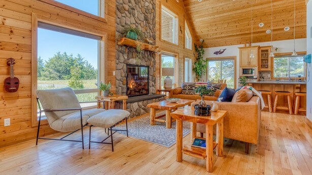 A spacious living area with a stone fireplace, wooden furniture, and large windows offering a view of the outdoors.