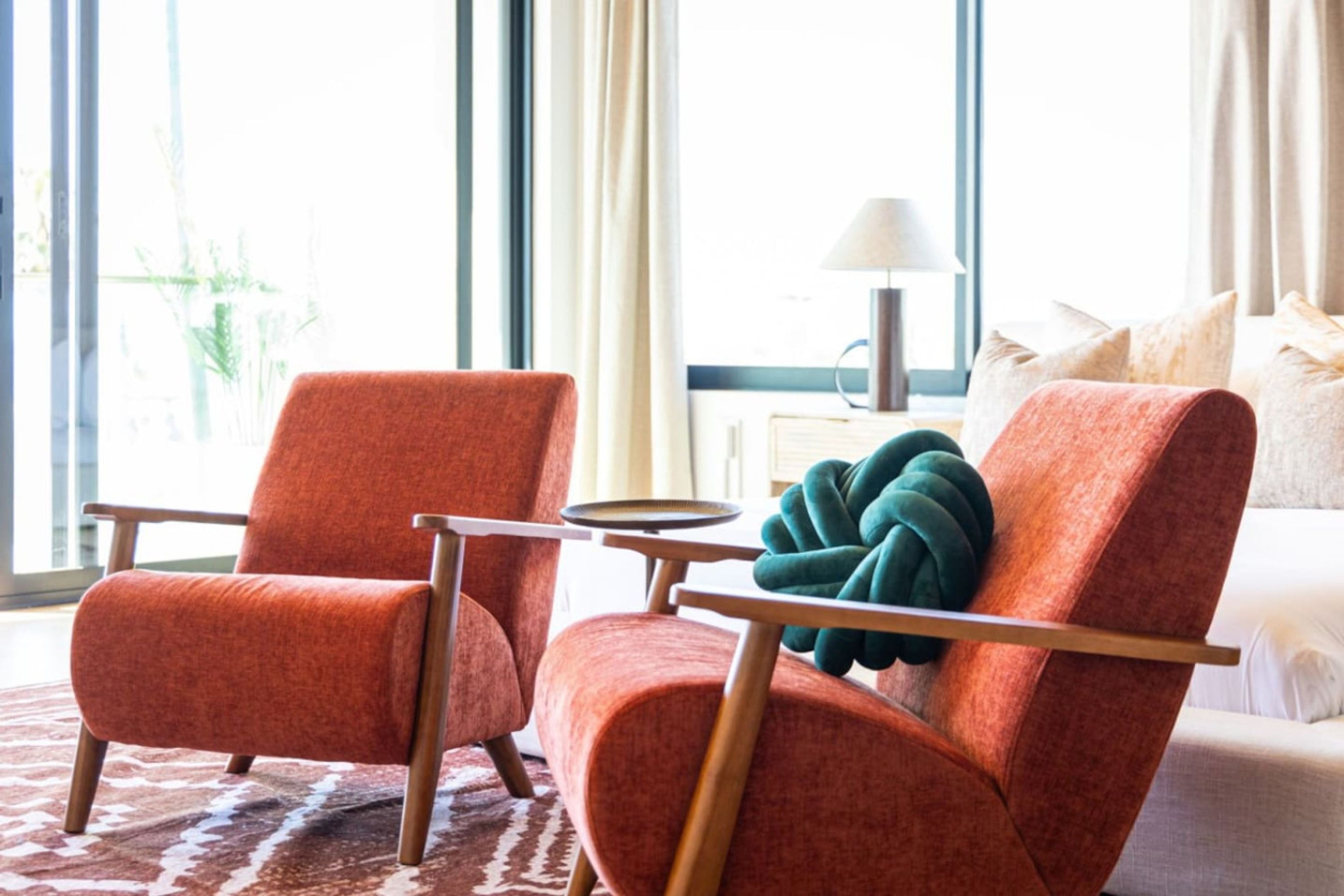 Una habitación de hotel moderna con dos sillones naranja, una mesita redonda, una manta verde y una cama con ropa blanca.
