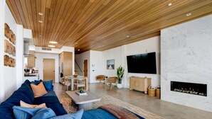 A modern living room with a wooden ceiling, a large TV, a fireplace, and a dining area.