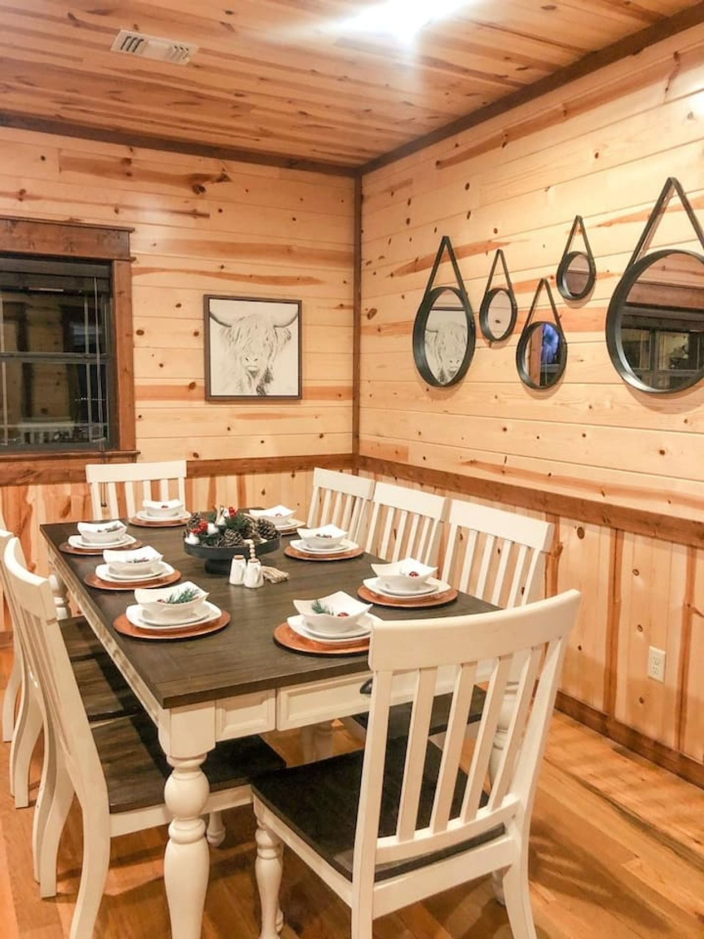 A wooden cabin dining room with a dark wooden table set for four, white chairs, and a framed animal picture on the wall.