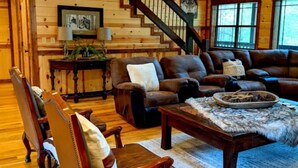 A spacious living room with a wooden staircase, leather sofas, and a large clock on the wall.