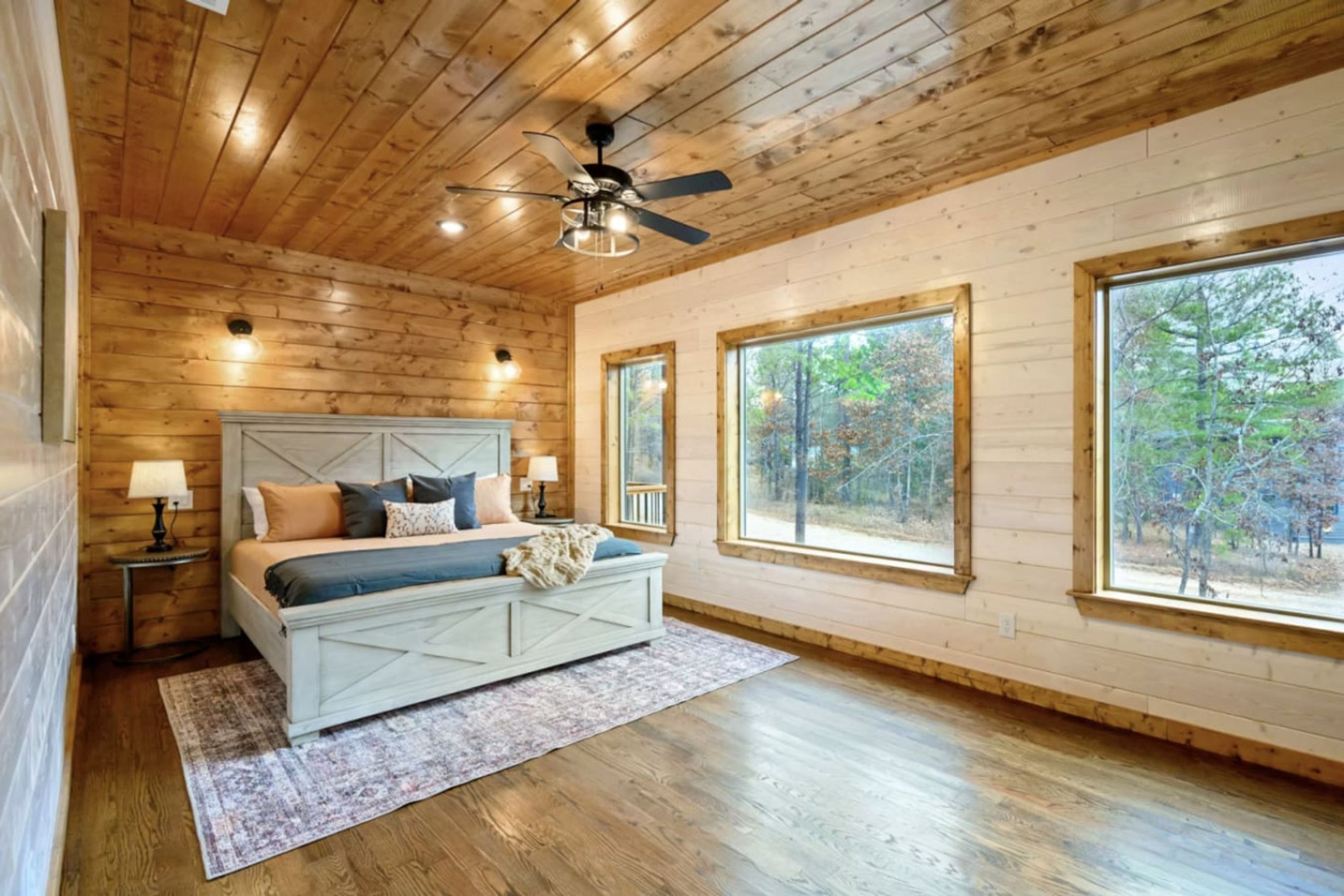 Un dormitorio con techo de madera, una cama con cabecera, dos mesitas de noche y ventanas grandes que dan a vistas de árboles.
