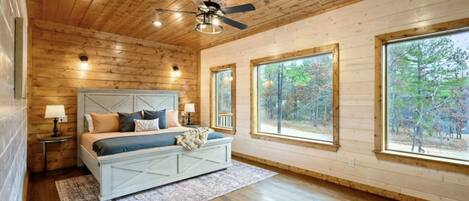 Un dormitorio con techo de madera, una cama con cabecera, dos mesitas de noche y ventanas grandes que dan a vistas de árboles.