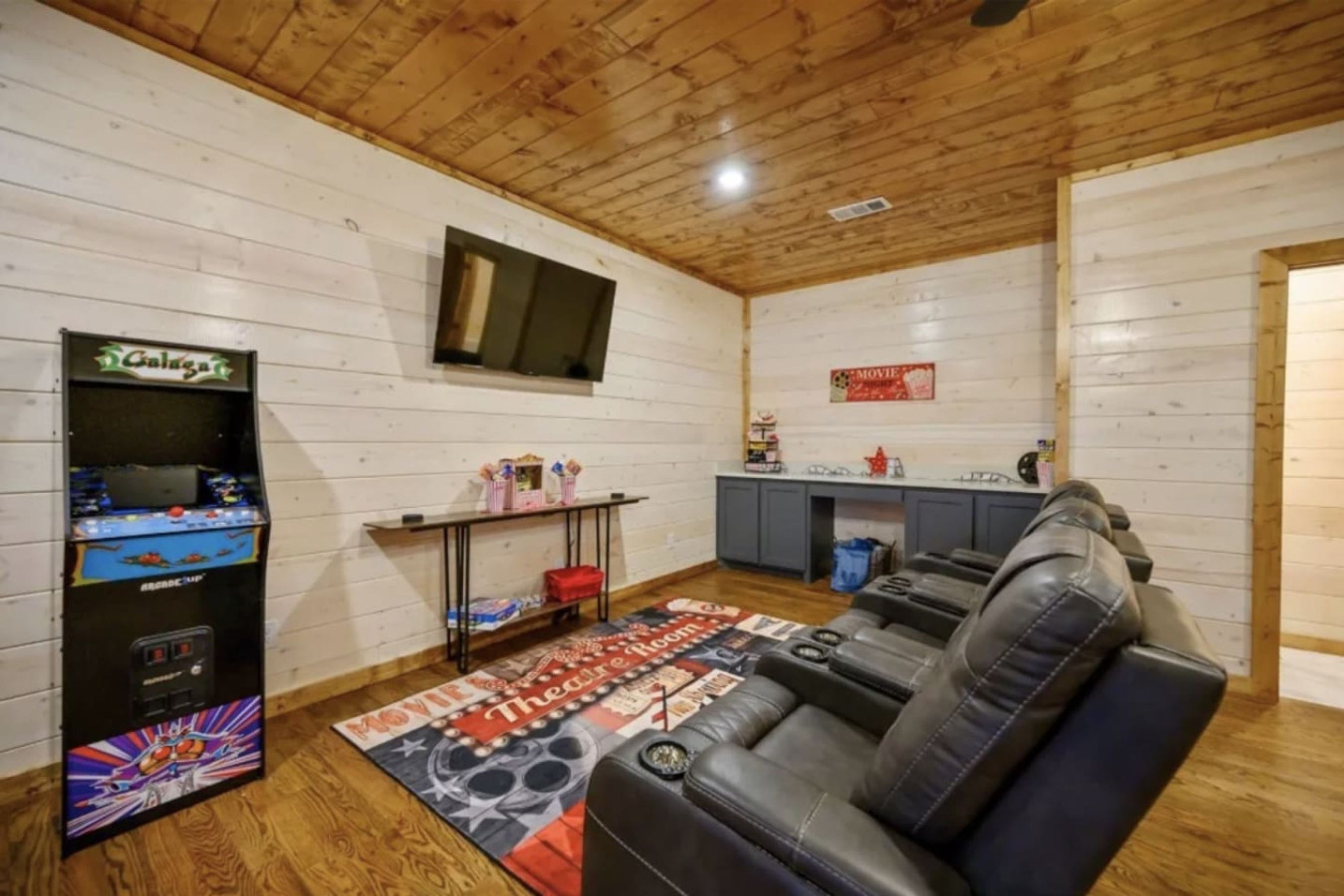 A cozy room with a wooden ceiling, a sofa, a gaming console, a kitchen area, and a TV mounted on the wall.