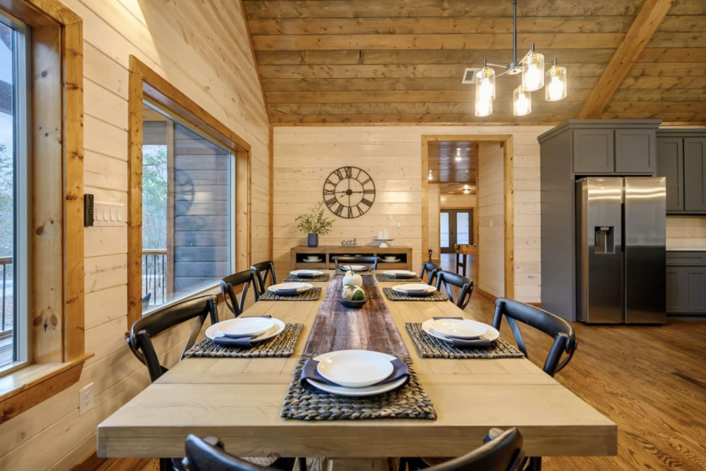 Una sala da pranzo in legno con un lungo tavolo apparecchiato per sei persone, una sveglia appesa al muro e un angolo cottura con frigorifero.