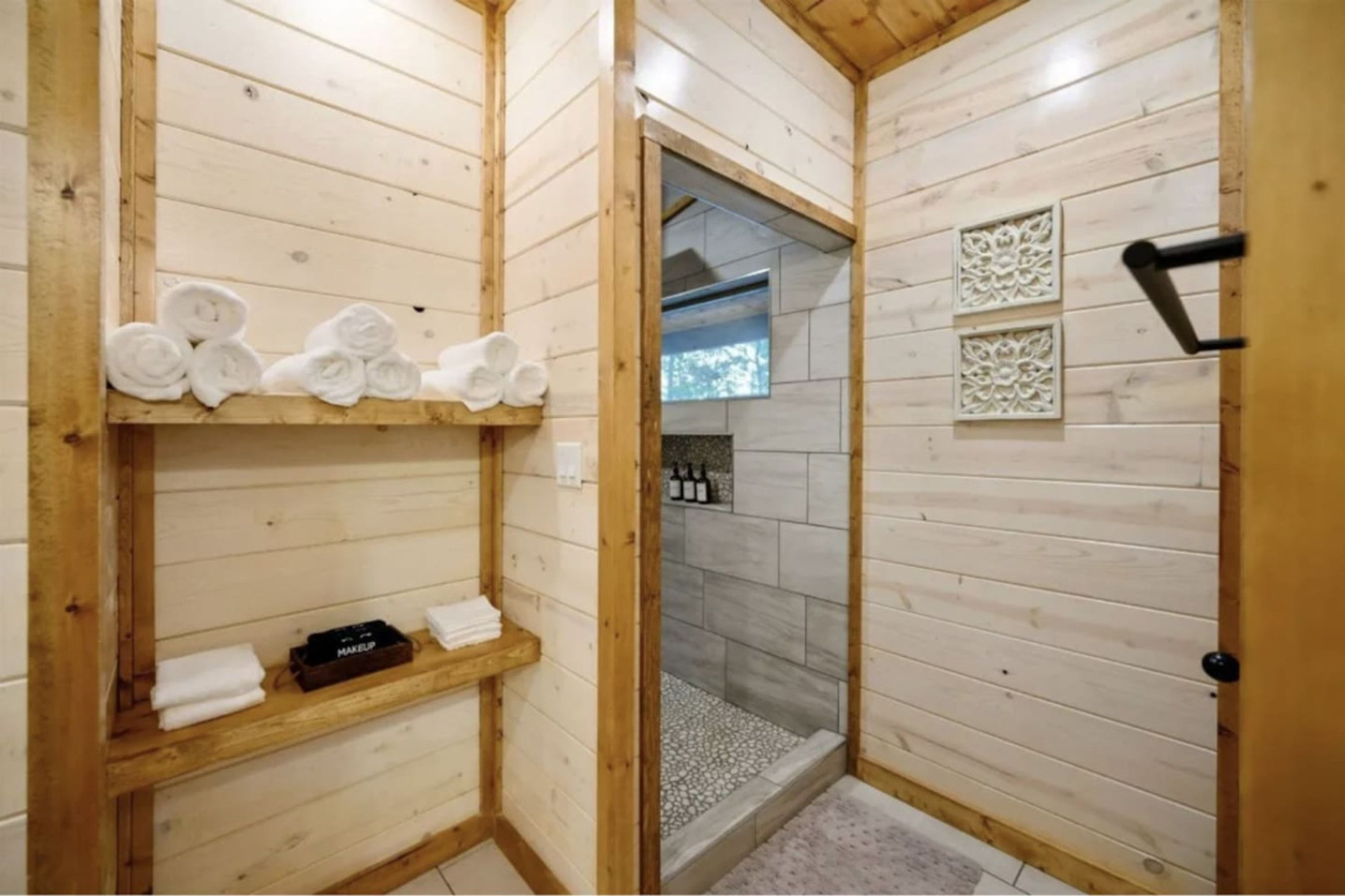 Une salle de bain rustique, aménagée dans une cabane en bois, avec une douche, des étagères où sont rangées les serviettes et une trousse de maquillage.