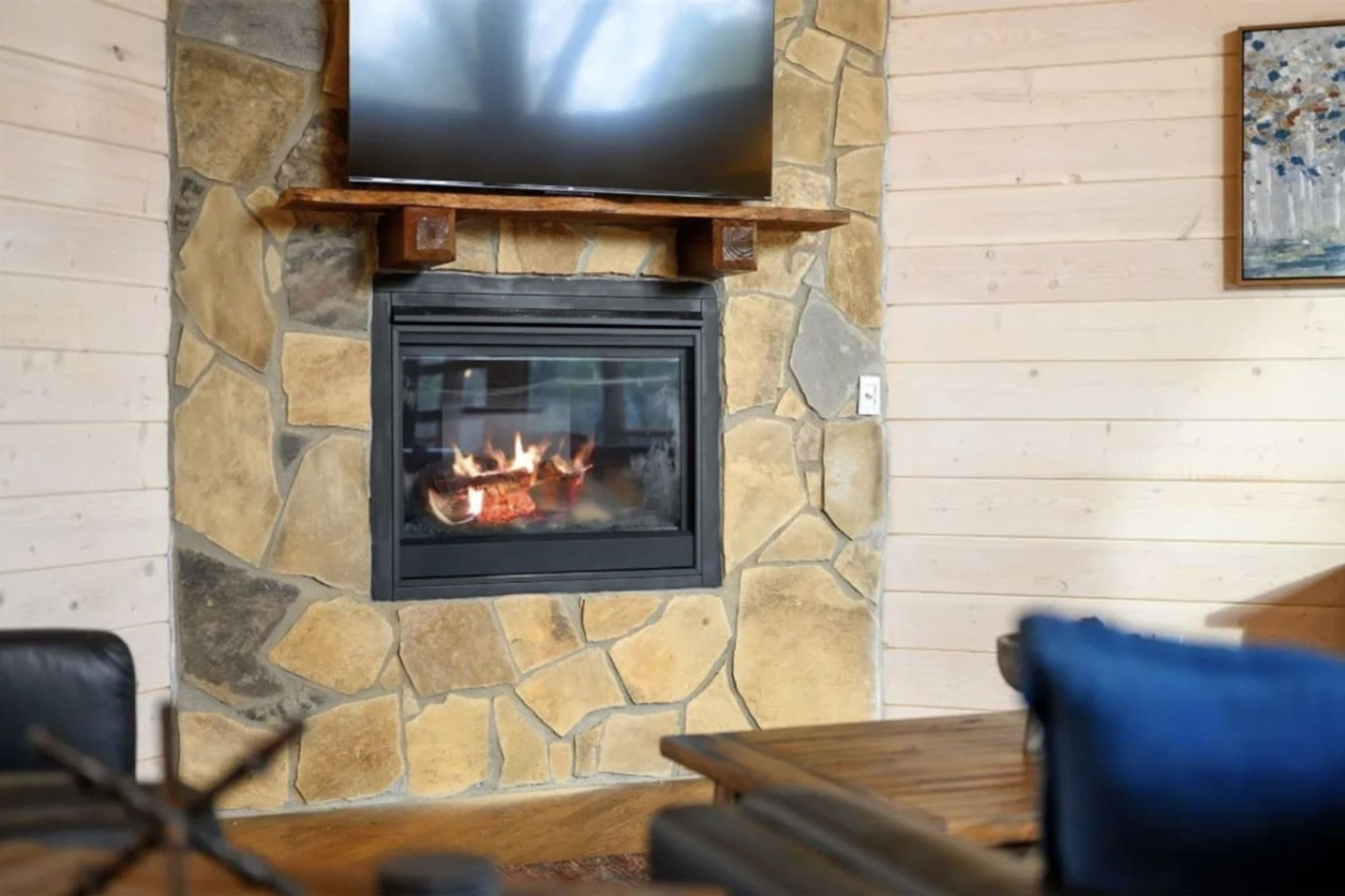 Ein Kamin aus Stein mit darüber montiertem Fernseher, ein Feuer brennt darin, und ein blaues Kissen auf einem Holztisch davor.