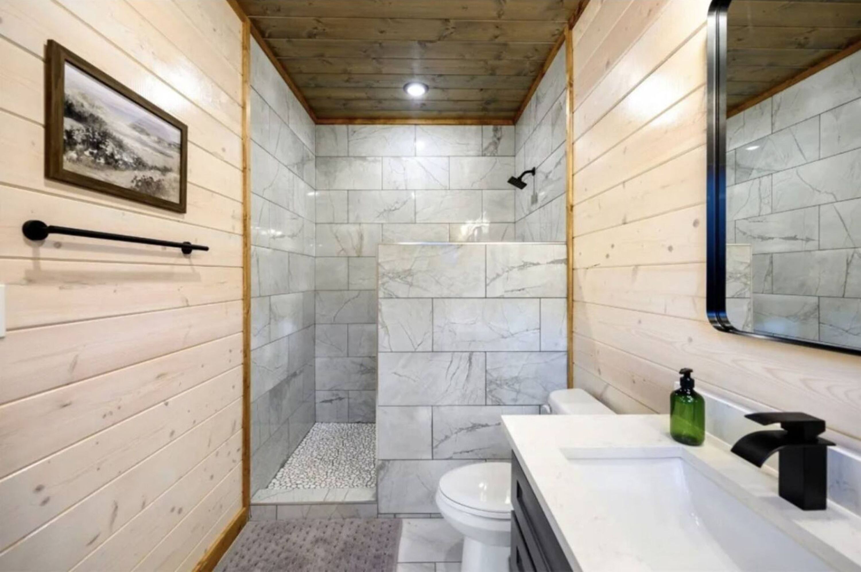 Ein Badezimmer mit Holzwänden, einer Marmordusche, einer Toilette und einem Waschbecken mit einem grünen Seifenspender.