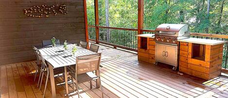A wooden deck with a table and chairs, a grill, and a view of trees.