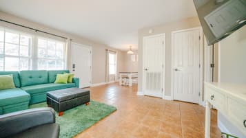 A living room with a green sofa, a coffee table, and a TV on a stand. There are two white doors and a window with blinds.