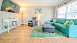 A living room with a flat-screen TV, a green sofa, a grey ottoman, a blue chair, and a small table with a lamp. - Convenient to Ole Miss The Square (Oxford)
